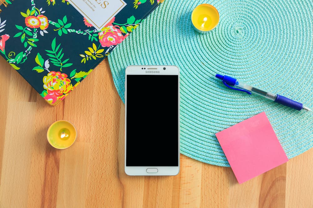 An old smartphone with a blank screen rests on a wood table next to an aqua round placemat, a pink sticky note, a floral notebook, and a blue pen. 